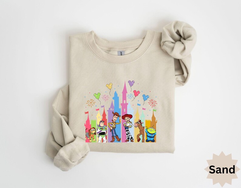 Peut inclure: Sweat-shirt couleur sable avec un graphisme color&eacute; des personnages de Toy Story devant un ch&acirc;teau avec des feux d'artifice et des ballons. Le sweat-shirt a des manches longues et un col rond. Le mot "Sand" est imprim&eacute; en bas &agrave; droite.