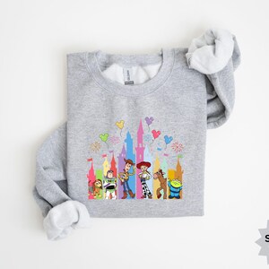 Peut inclure: Un sweat-shirt gris clair avec un motif color&eacute; de personnages de dessins anim&eacute;s et une silhouette de ch&acirc;teau. Le motif comprend des personnages comme Woody, Buzz l'&Eacute;clair et d'autres, avec des ballons et des feux d'artifice. Le sweat-shirt est &eacute;tiquet&eacute; "Sports Grey".