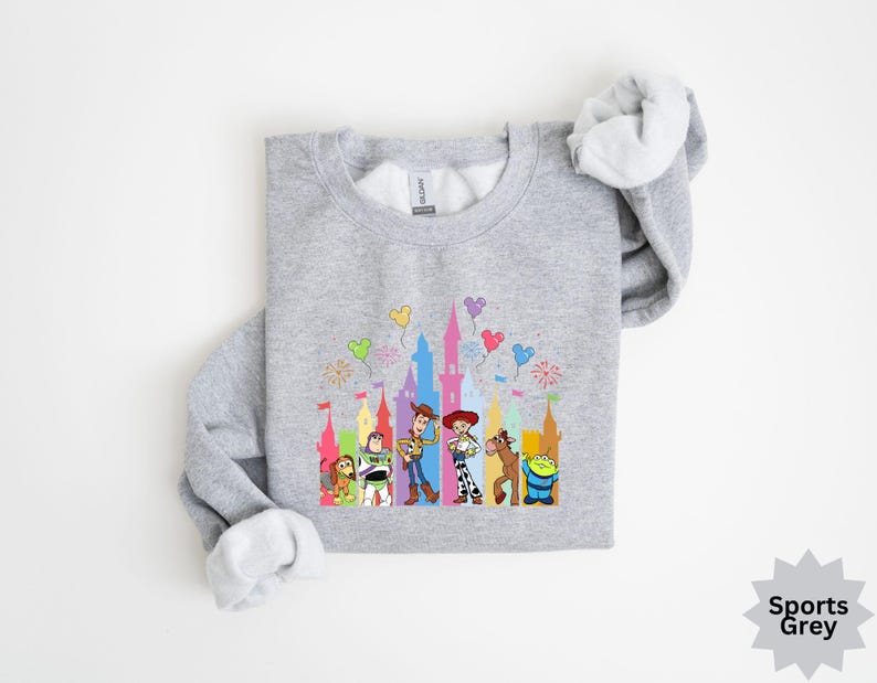 Peut inclure: Un sweat-shirt gris clair avec un motif color&eacute; de personnages de dessins anim&eacute;s et une silhouette de ch&acirc;teau. Le motif comprend des personnages comme Woody, Buzz l'&Eacute;clair et d'autres, avec des ballons et des feux d'artifice. Le sweat-shirt est &eacute;tiquet&eacute; "Sports Grey".
