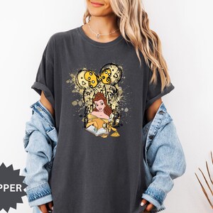 Puede incluir: Camiseta gris oscuro con un dise&ntilde;o de Bella de La Bella y la Bestia leyendo un libro, con un dise&ntilde;o inspirado en Mickey Mouse en oro y amarillo. La camiseta tiene un corte holgado y mangas cortas.