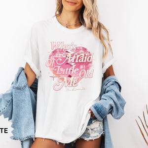 May include: White t-shirt with a pink graphic of a disco ball and the text "Who's Afraid of Little Old Me". The shirt is a relaxed fit and has short sleeves. A light blue denim jacket is draped over the shoulders.