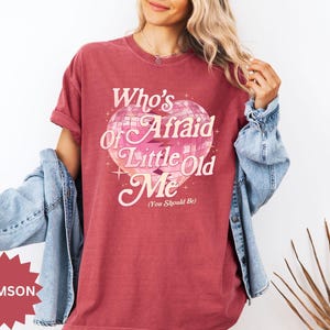 May include: Crimson-colored t-shirt with the text "Who's Afraid of Little Old Me (You Should Be)" in a retro font. The graphic features a disco ball heart design with pink and white accents. A denim jacket is draped over the shoulders.