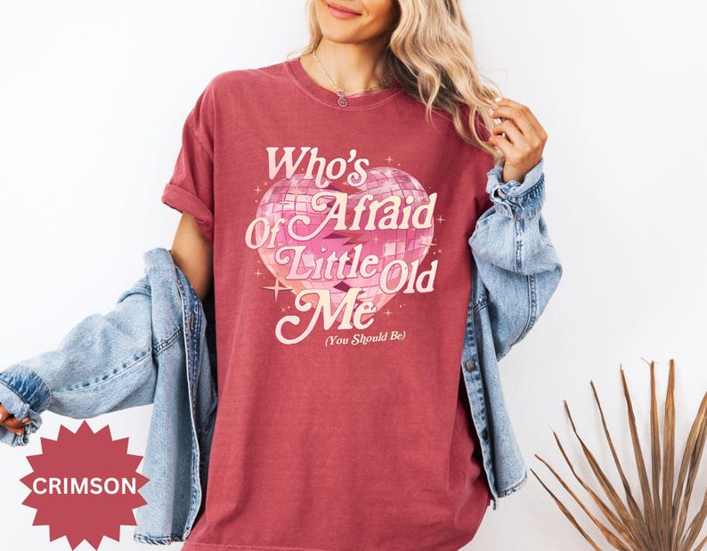 May include: Crimson-colored t-shirt with the text "Who's Afraid of Little Old Me (You Should Be)" in a retro font. The graphic features a disco ball heart design with pink and white accents. A denim jacket is draped over the shoulders.