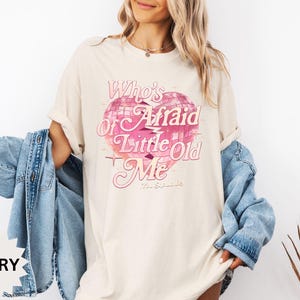 May include: Ivory-colored t-shirt with a pink heart-shaped graphic and the text "Who's Afraid of Little Old Me". The shirt has a relaxed fit and rolled-up sleeves. A light blue denim jacket is draped over the shoulders.