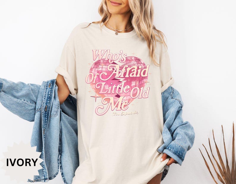 May include: Ivory-colored t-shirt with a pink heart-shaped graphic and the text "Who's Afraid of Little Old Me". The shirt has a relaxed fit and rolled-up sleeves. A light blue denim jacket is draped over the shoulders.