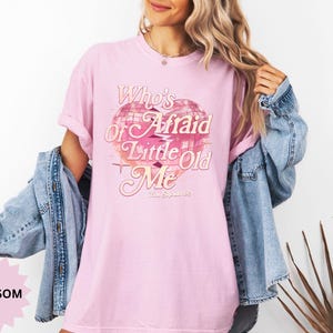 May include: A light pink t-shirt with the text "Who's Afraid of Little Old Me" in a vintage style. The graphic includes a pink and white disco ball design. The shirt is paired with a denim jacket.