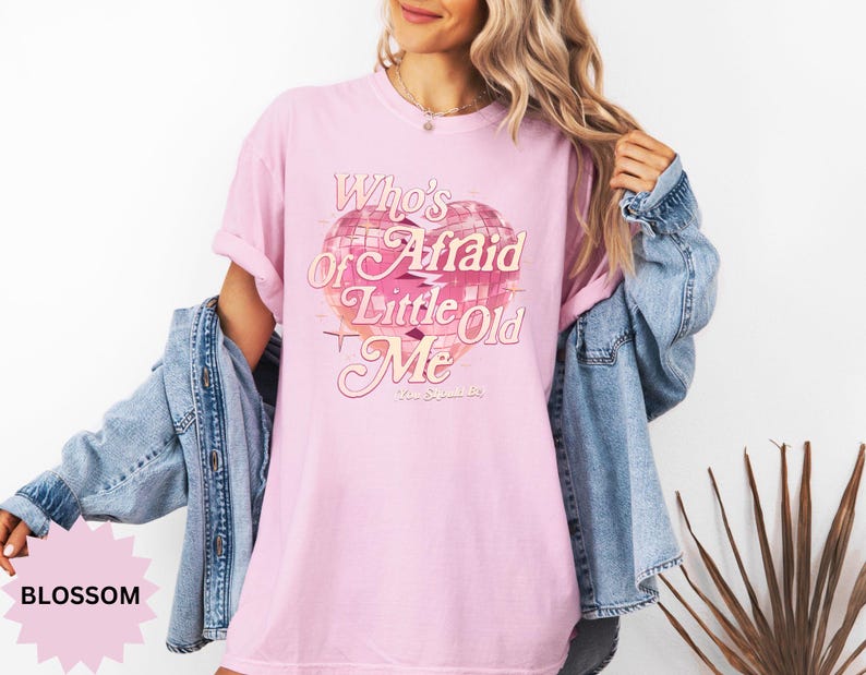 May include: A light pink t-shirt with the text "Who's Afraid of Little Old Me" in a vintage style. The graphic includes a pink and white disco ball design. The shirt is paired with a denim jacket.