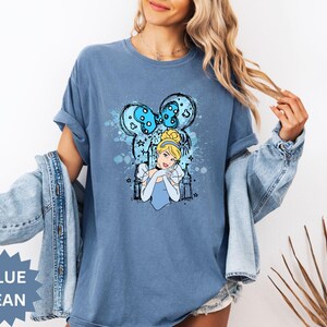 Puede incluir: Camiseta azul vaquero con un gr&aacute;fico de Cenicienta. El dise&ntilde;o incluye una imagen de dibujos animados de Cenicienta, un lazo azul y estrellas. Las palabras "BLUE JEAN" est&aacute;n impresas en una forma de estrella azul.