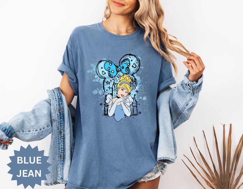Puede incluir: Camiseta azul vaquero con un gr&aacute;fico de Cenicienta. El dise&ntilde;o incluye una imagen de dibujos animados de Cenicienta, un lazo azul y estrellas. Las palabras "BLUE JEAN" est&aacute;n impresas en una forma de estrella azul.