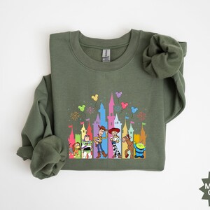 Peut inclure: Sweat-shirt vert militaire &agrave; col rond avec un graphisme color&eacute; de personnages de dessins anim&eacute;s devant un ch&acirc;teau. Le graphisme comprend des personnages comme Woody, Buzz l'&Eacute;clair, etc. Le sweat-shirt est en mati&egrave;re douce.