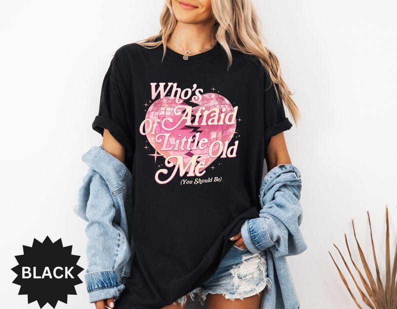 May include: Black t-shirt with the text "Who's Afraid of Little Old Me" in pink script and a pink heart-shaped disco ball graphic. The shirt has short sleeves and a relaxed fit. The word "BLACK" is in a black starburst.