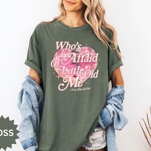 May include: A moss green t-shirt with the text "Who's Afraid of Little Old Me" in pink, featuring a disco ball heart design. The shirt has short sleeves and a relaxed fit.