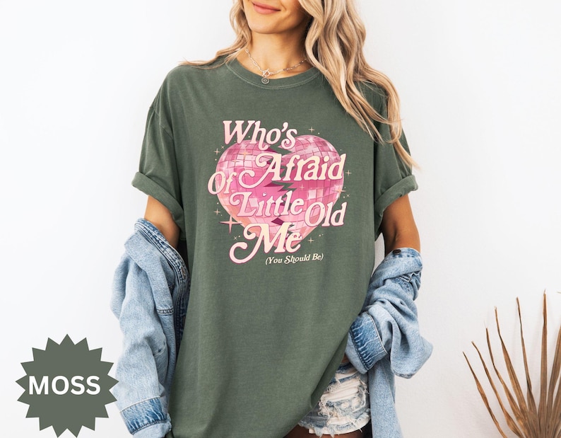May include: A moss green t-shirt with the text "Who's Afraid of Little Old Me" in pink, featuring a disco ball heart design. The shirt has short sleeves and a relaxed fit.