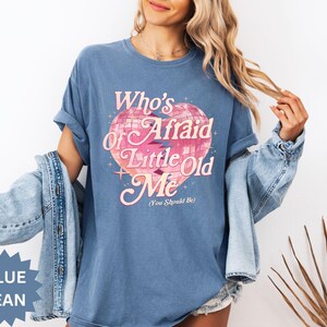 May include: A blue jean-colored t-shirt with the text "Who's Afraid of Little Old Me (You Should Be)" in pink and white. The graphic features a pink heart-shaped disco ball design. The shirt is a relaxed fit.