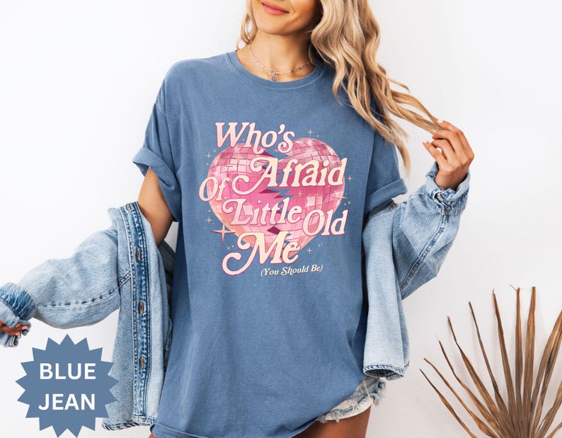 May include: A blue jean-colored t-shirt with the text "Who's Afraid of Little Old Me (You Should Be)" in pink and white. The graphic features a pink heart-shaped disco ball design. The shirt is a relaxed fit.