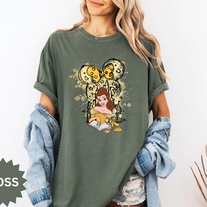Puede incluir: Camiseta verde musgo con un gr&aacute;fico de Bella de La Bella y la Bestia, leyendo un libro. El dise&ntilde;o incluye una silueta de oreja de Mickey Mouse con detalles amarillos y estrellas. La camiseta es de corte holgado.
