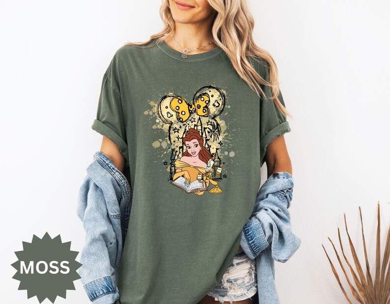 Puede incluir: Camiseta verde musgo con un gr&aacute;fico de Bella de La Bella y la Bestia, leyendo un libro. El dise&ntilde;o incluye una silueta de oreja de Mickey Mouse con detalles amarillos y estrellas. La camiseta es de corte holgado.