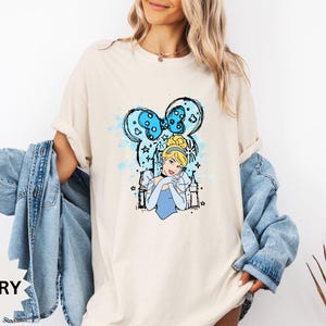 Puede incluir: Camiseta color marfil con un dise&ntilde;o de Cenicienta. El dise&ntilde;o incluye un lazo azul, el retrato de Cenicienta e ilustraciones de castillos. La camiseta tiene un corte holgado y mangas cortas.