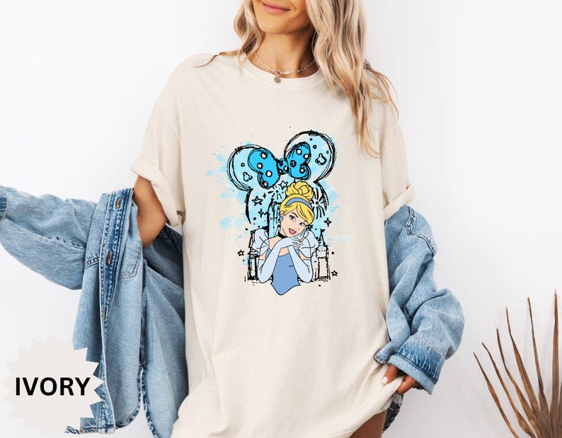 Puede incluir: Camiseta color marfil con un dise&ntilde;o de Cenicienta. El dise&ntilde;o incluye un lazo azul, el retrato de Cenicienta e ilustraciones de castillos. La camiseta tiene un corte holgado y mangas cortas.