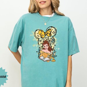 Puede incluir: Camiseta verde mar con un dise&ntilde;o de Bella de La Bella y la Bestia, con un dise&ntilde;o inspirado en Mickey Mouse. El dise&ntilde;o incluye a Bella leyendo un libro, con detalles en amarillo y negro. La camiseta tiene un corte c&oacute;modo y holgado.