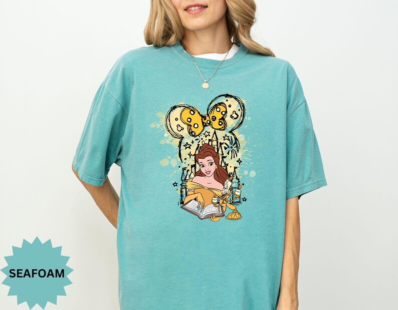 Puede incluir: Camiseta verde mar con un dise&ntilde;o de Bella de La Bella y la Bestia, con un dise&ntilde;o inspirado en Mickey Mouse. El dise&ntilde;o incluye a Bella leyendo un libro, con detalles en amarillo y negro. La camiseta tiene un corte c&oacute;modo y holgado.