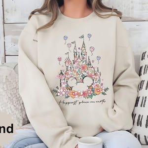May include: A beige sweatshirt with a floral design of a castle and Mickey Mouse ears. The text "Happiest place on earth" is printed below the design.