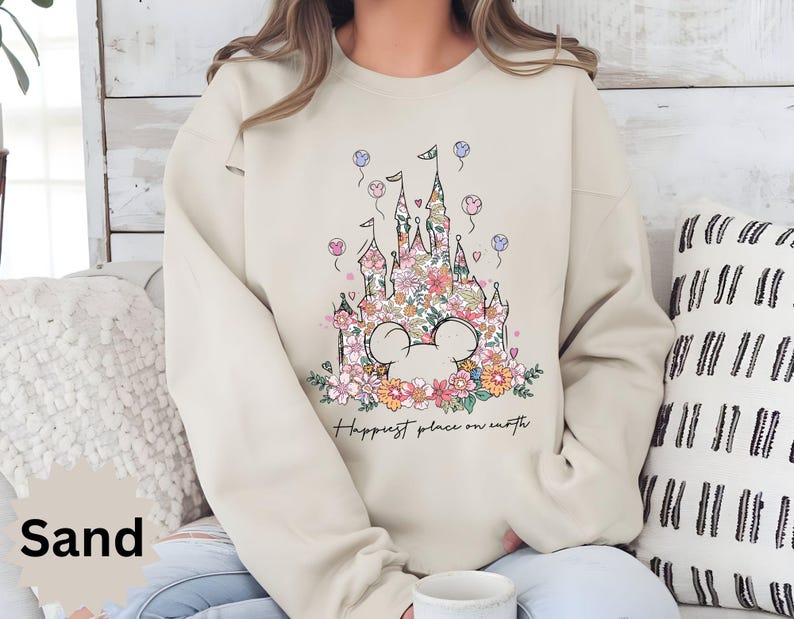 May include: A beige sweatshirt with a floral design of a castle and Mickey Mouse ears. The text "Happiest place on earth" is printed below the design.