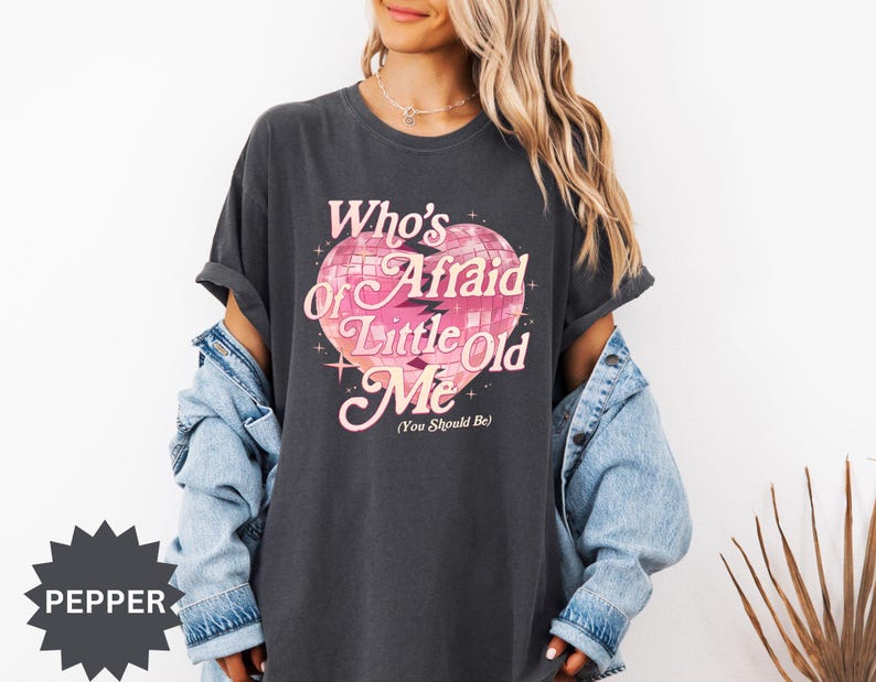 May include: Dark gray t-shirt with the text "Who's Afraid of Little Old Me (You Should Be)" in pink and white. The graphic is a heart-shaped disco ball. The shirt has short sleeves and a relaxed fit.