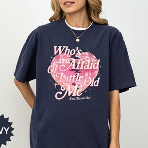 May include: Navy blue t-shirt with the text "Who's Afraid of Little Old Me" in pink and white, featuring a disco ball heart design. The shirt has short sleeves and a round neckline.