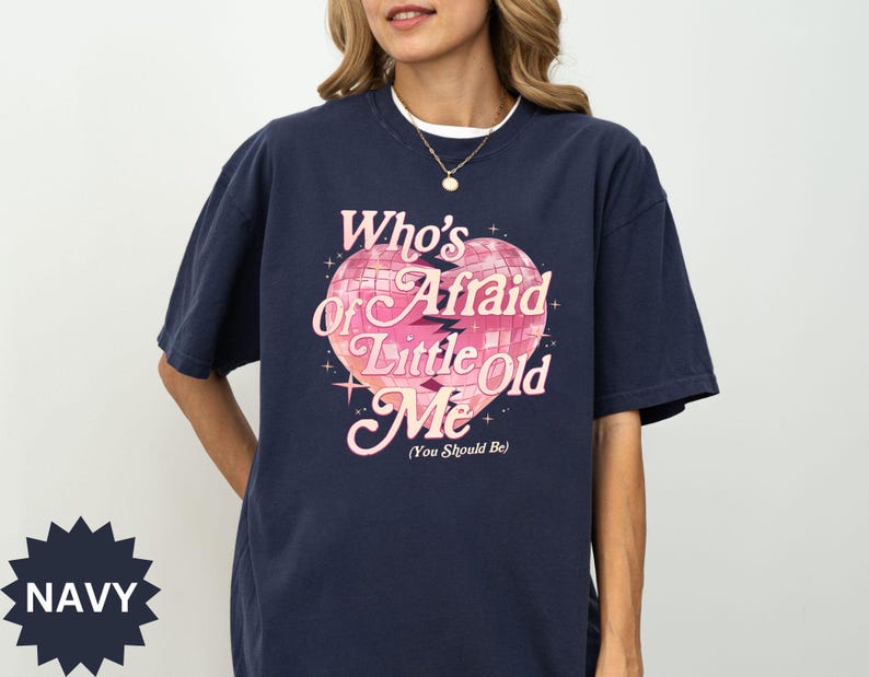 May include: Navy blue t-shirt with the text "Who's Afraid of Little Old Me" in pink and white, featuring a disco ball heart design. The shirt has short sleeves and a round neckline.