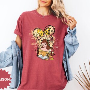 Puede incluir: Camiseta carmes&iacute; con una ilustraci&oacute;n de dibujos animados de Bella de La Bella y la Bestia leyendo un libro. El dise&ntilde;o incluye una cabeza inspirada en Mickey Mouse con detalles amarillos y estrellas.