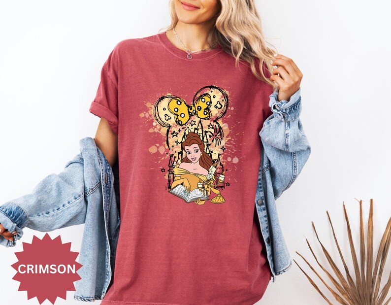 Puede incluir: Camiseta carmes&iacute; con una ilustraci&oacute;n de dibujos animados de Bella de La Bella y la Bestia leyendo un libro. El dise&ntilde;o incluye una cabeza inspirada en Mickey Mouse con detalles amarillos y estrellas.