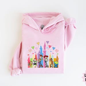 Peut inclure: Sweat &agrave; capuche rose clair avec un motif color&eacute; d'un ch&acirc;teau et de personnages de dessins anim&eacute;s. Le motif comprend des personnages comme Woody et Buzz l'&Eacute;clair. Le sweat &agrave; capuche a une capuche &agrave; cordon et des manches longues. Le texte "Light Pink" est en bas &agrave; droite.