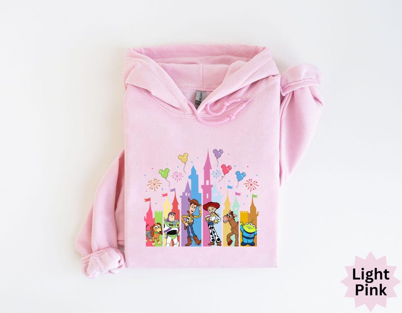 Peut inclure: Sweat &agrave; capuche rose clair avec un motif color&eacute; d'un ch&acirc;teau et de personnages de dessins anim&eacute;s. Le motif comprend des personnages comme Woody et Buzz l'&Eacute;clair. Le sweat &agrave; capuche a une capuche &agrave; cordon et des manches longues. Le texte "Light Pink" est en bas &agrave; droite.
