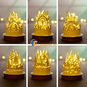 May include: A collection of illuminated paper cutouts depicting Christmas scenes. Each cutout is a different design, including a nativity scene, a forest with deer, and a castle with the text "Merry Christmas". The cutouts sit on wooden bases.