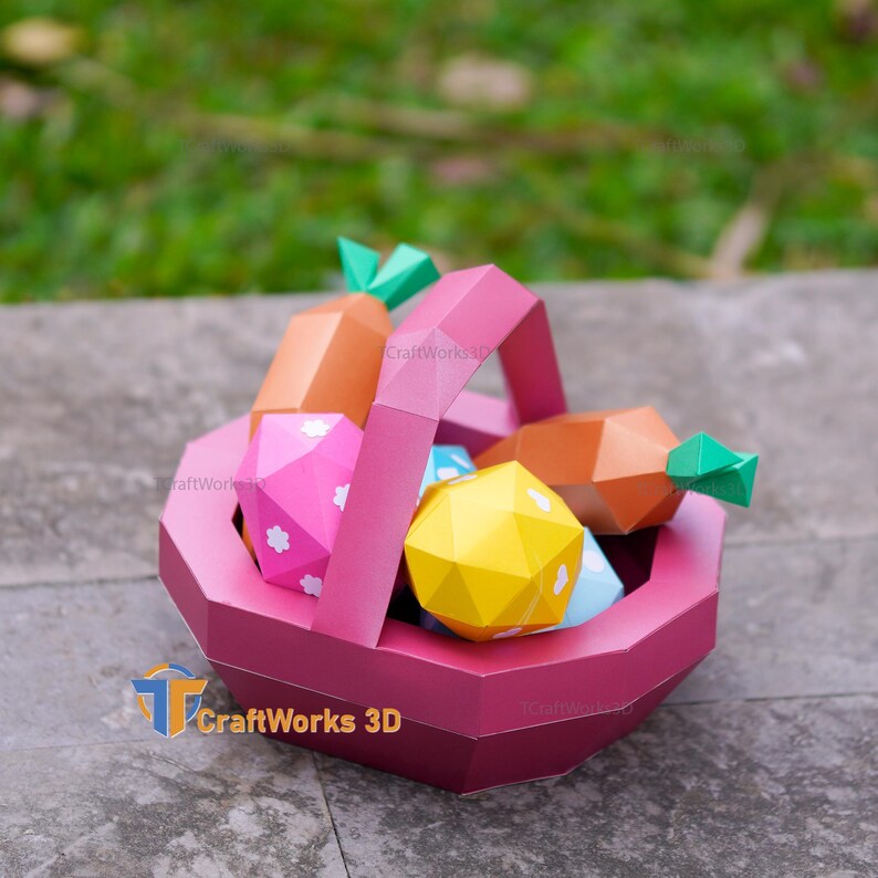 K&ouml;nnte beinhalten: Ein Papier-Osterkorb in Rosat&ouml;nen, gef&uuml;llt mit geometrischen Papiereiern und Karotten. Der Korb hat einen Henkel und steht auf einer Steinoberfl&auml;che. Korb und Inhalt bestehen aus gefaltetem Papier.
