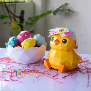 Può includere: Un pulcino di carta giallo con una corona floreale è seduto accanto a un guscio d'uovo di carta bianco pieno di colorate uova di Pasqua geometriche. Le uova sono blu, rosa e gialle con pois bianchi. Ci sono scaglie di carta rossa sparse.