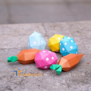 K&ouml;nnte beinhalten: Eine Sammlung farbenfroher, geometrischer Papier-Gem&uuml;se- und Ostereiformen. Die orangefarbenen Karotten haben gr&uuml;ne Spitzen, und die Eier sind blau, gelb und rosa mit wei&szlig;en Tupfen und Blumenakzenten. Der Text "TCraftWorks3D" ist sichtbar.