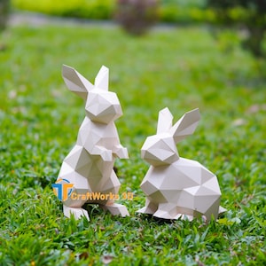 Pode incluir: Duas esculturas de coelhos geométricos em papel branco sujo, uma em pé e outra sentada, são exibidas sobre relva verde. As esculturas têm um design poligonal moderno. O logotipo "CraftWorks 3D" é visível.