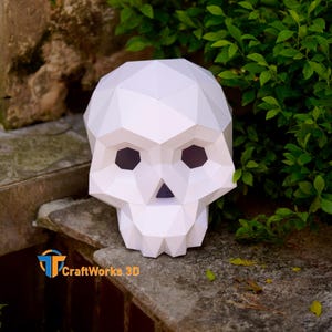 May include: A white, geometric paper skull sculpture with a polygonal design. The skull has dark eye sockets and a triangular nose. The sculpture is displayed outdoors, with green foliage in the background. The CraftWorks 3D logo is visible.