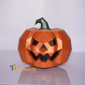 May include: A geometric orange pumpkin with a carved face and a green stem. The pumpkin is made of folded paper and has a 3D effect. The CraftWorks 3D logo is visible in the lower left corner. A Halloween decoration.