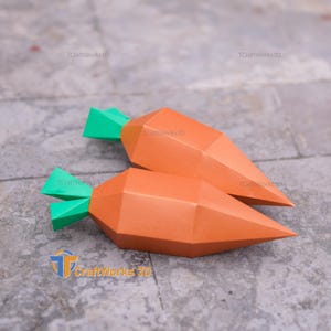 K&ouml;nnte beinhalten: Zwei orangefarbene Papierkarotten mit gr&uuml;nen Bl&auml;ttern liegen auf einer grauen Oberfl&auml;che. Die Karotten haben ein geometrisches, facettiertes Design. Das Logo "TCraftWorks3D" ist sichtbar.
