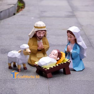 May include: A low-poly paper craft nativity scene featuring a figure in a tan robe, a figure in a blue robe with a white head covering, a small infant in a manger, and a white sheep. The figures have geometric facets and the text CraftWorks 3D.