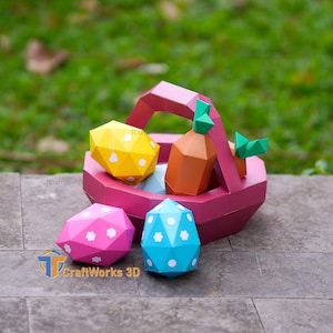 Könnte beinhalten: Ein rosa Papier-Osterkorb gefüllt mit einem gelben Ei, einer orangefarbenen Karotte und blauen und rosa Eiern. Die Eier und die Karotte haben weiße Blumenmuster. Der Korb steht auf einer grauen Steinoberfläche.