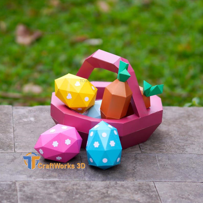 K&ouml;nnte beinhalten: Ein rosa Papier-Osterkorb gef&uuml;llt mit einem gelben Ei, einer orangefarbenen Karotte und blauen und rosa Eiern. Die Eier und die Karotte haben wei&szlig;e Blumenmuster. Der Korb steht auf einer grauen Steinoberfl&auml;che.