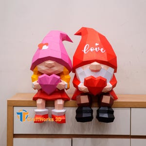 May include: Two papercraft gnomes, one female and one male, holding heart-shaped objects. The female gnome wears a pink hat and dress, while the male gnome has a red hat with the word "love" on it. The gnomes are sitting on a wooden surface.