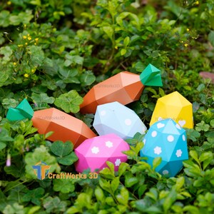 K&ouml;nnte beinhalten: Eine Sammlung farbenfroher, geometrischer Osterdekorationen aus Papier, darunter orangefarbene Karotten mit gr&uuml;nen Spitzen und pastellfarbene Eier mit wei&szlig;en Tupfen und Blumenmustern. Die Artikel sind in gr&uuml;nem Laub eingebettet.