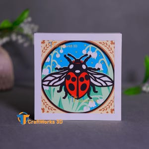 Pode incluir: Um cartão branco com uma ilustração colorida de uma joaninha com asas pretas e vermelhas e um corpo vermelho com manchas pretas. A joaninha está cercada por folhas verdes e flores brancas. O cartão tem uma borda marrom com um design floral.