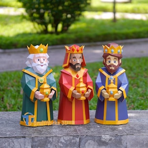 Puede incluir: Tres figuras de papel low-poly que representan a los Reyes Magos, cada una con corona y sosteniendo un recipiente dorado. Las figuras visten túnicas verdes, rojas y moradas con adornos amarillos, caracterizadas por facetas geométricas.