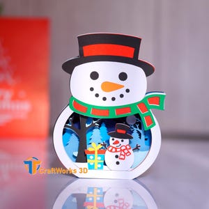 May include: A layered paper snowman decoration with a black top hat, orange nose, and green scarf. Inside the snowman is a winter scene with a smaller snowman, trees, and a gift. The base has the text "CraftWorks 3D".